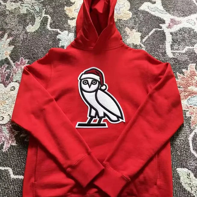 Red OVO Christmas hoodie with OG Owl logo wearing Santa hat on patterned rug