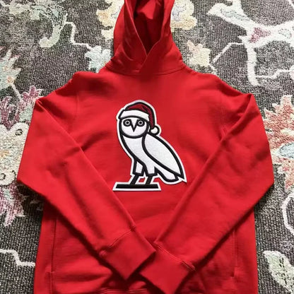 Red OVO Christmas hoodie with OG Owl logo wearing Santa hat on patterned rug